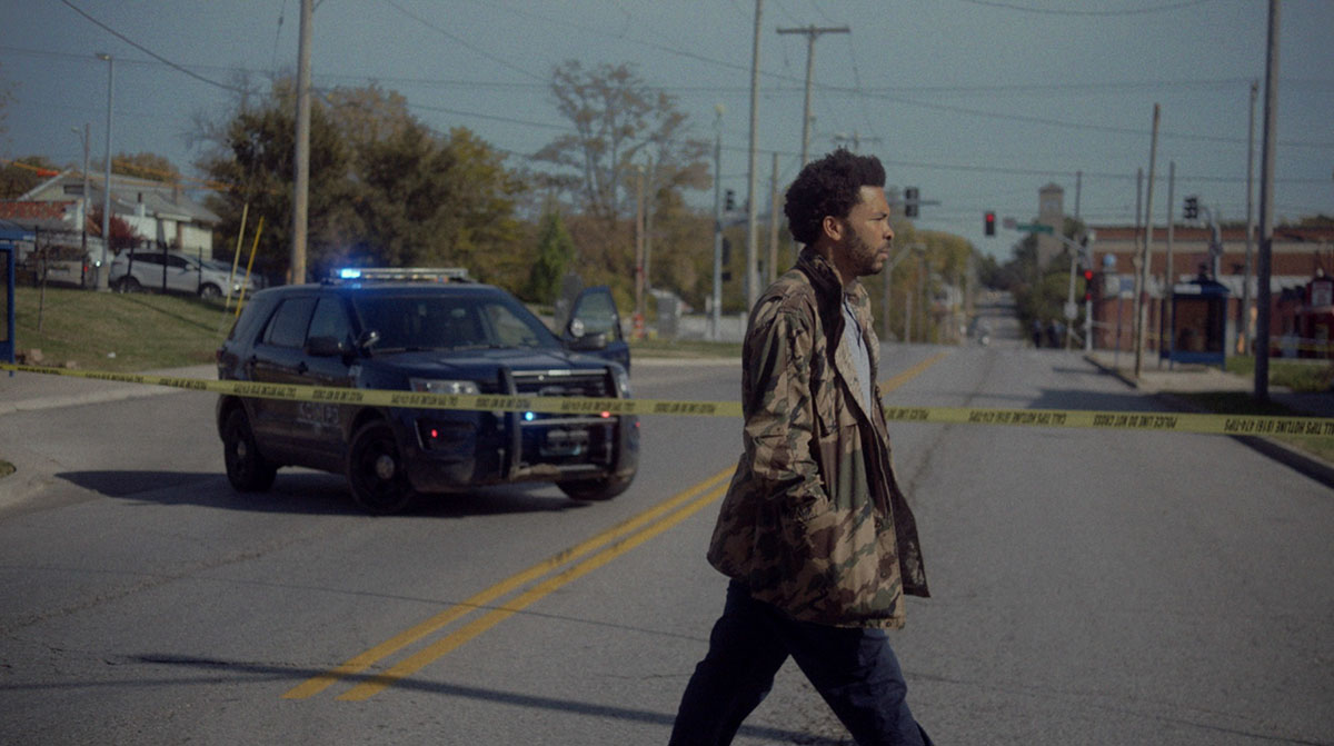 Artist George Morton is a Black man seen crossing a street demarcated by police tape. He is wearing dark pants and a camo jacket. From Rosa Ruth Boesten’s ‘Master of Light,’ which won the McBaine Documentary Feature Award at SFFILM 2022. Courtesy of SFFILM.