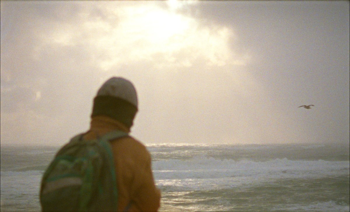 A person wearing an orange jacket and a white cap looks out at the sea. From Jacquelyn Mills' 'Geographies of Solitude.' Courtesy of Hot Docs.