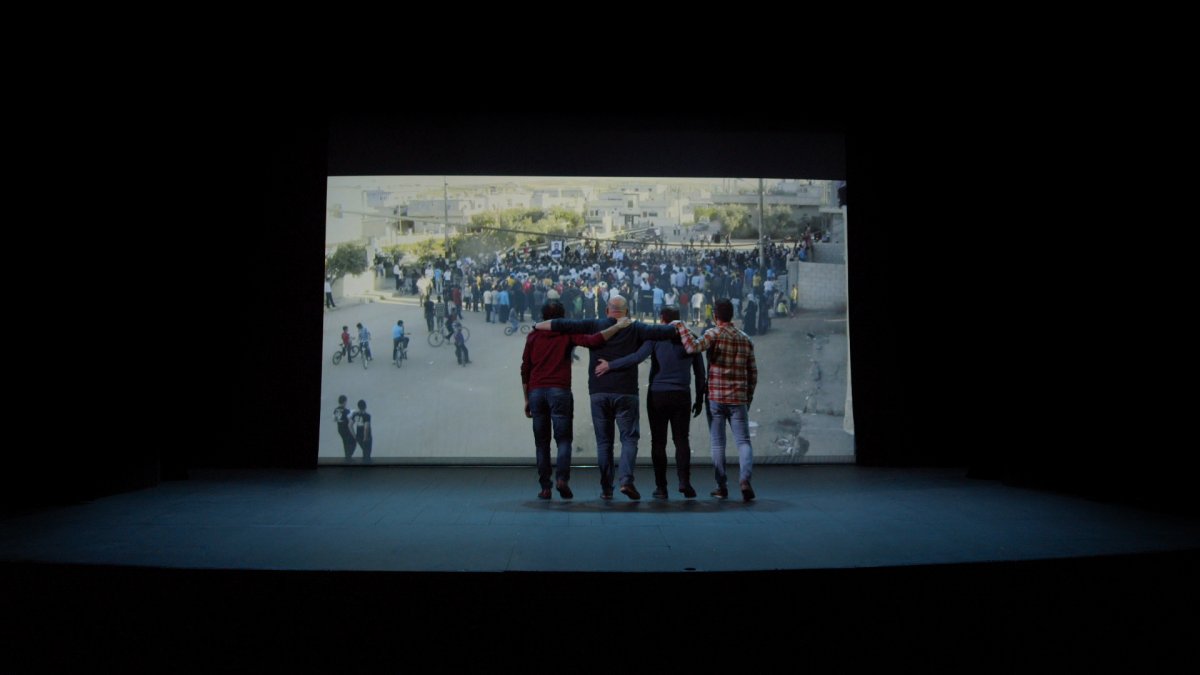 Four men doing the Dabkha dance in front of a screen showing their footage of the Syrian revolution. From Rami Farah's 'Our Memory Belongs to Us.' Courtesy of Hot Docs.