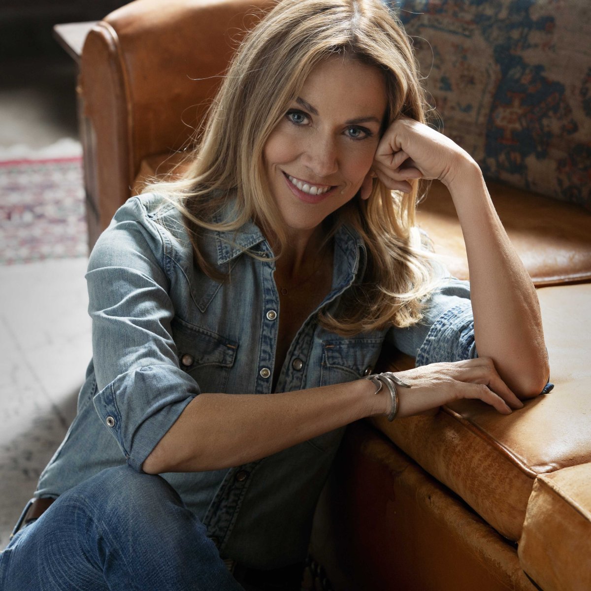 American musician Sheryl Crow is a white woman with blonde hair wearing a button-down shirt, sitting on the floor and smiling toward the camera. 