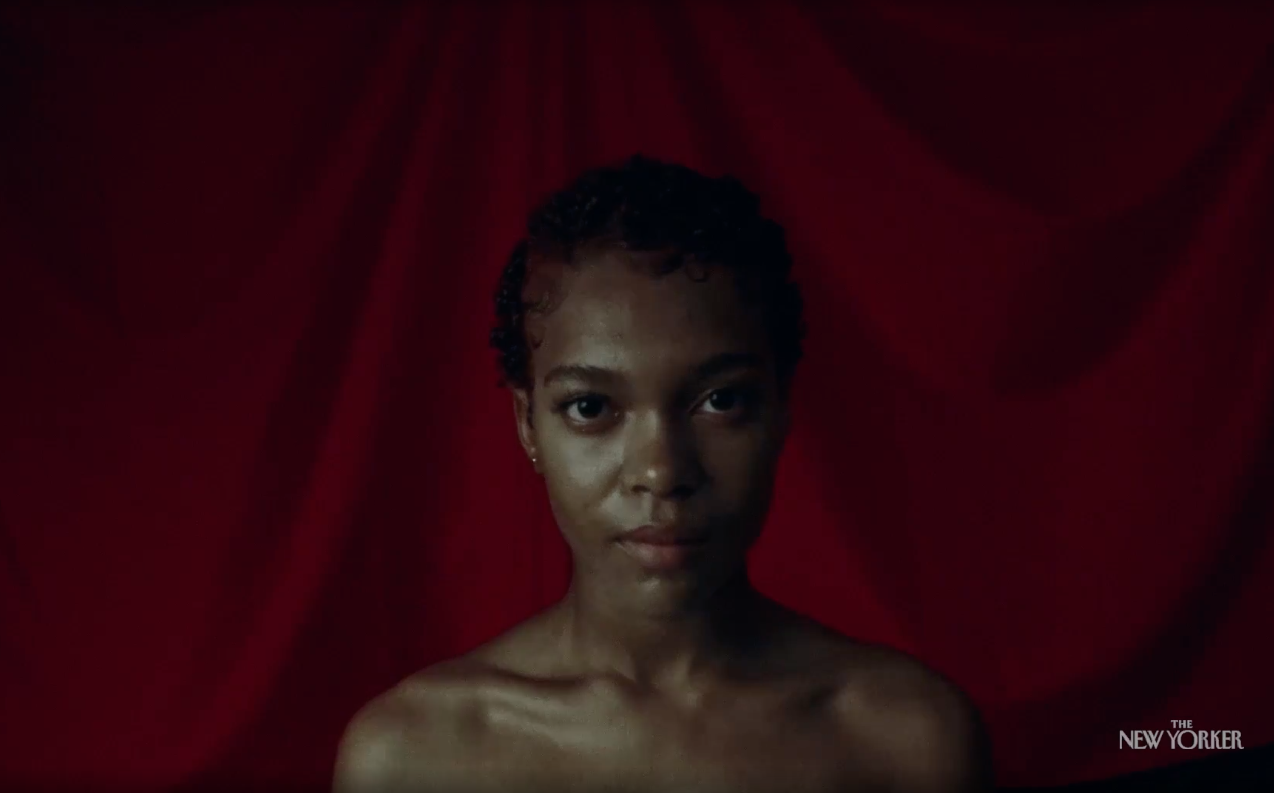 A Black woman with short curly hair stands in front of a red background, bare-shouldered and staring at the camera. From Juliana Kasumu’s 'Baby Bangz: Black Power in Hair,' now streaming on New Yorker.com.