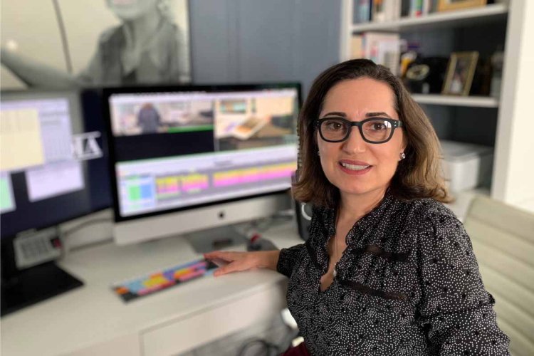 Editor Darianna Cardilli, a white woman with glasses, brown, shoulder-length hair and a patterned blouse, poses in front of her editing bay. Courtesy of Darianna Cardilli.