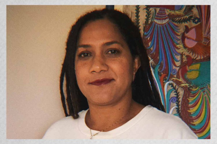 Polaroid headshot of Sonya Childress, a Black woman wearing a white t-shirt and a silver necklace, standing in front of a wall with a colorful painting.