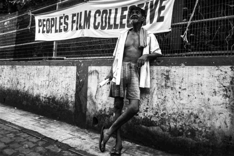 An elderly Indian man stands on a sidewalk, wearing knee-length shorts and an unbuttoned shirt. He is leaning against a People's Film Collective banner. Photo courtesy of People's Film Collective.