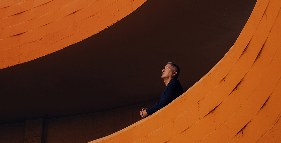 Richard Ray "Rick" Perez is a Mexican-American man with short greying hair, wearing a dark blue blazer, standing behind a patterned orange wall and looking up.