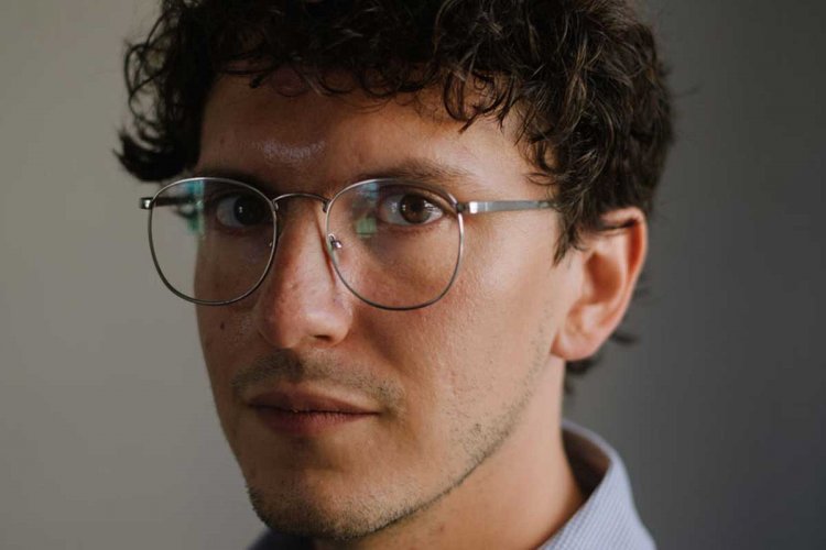 A portrait shot of Gerardo del Valle in front of a gray wall. He is wearing a dress shirt, wears glasses, and has short curly brown hair. (Photo by Gili Benita)