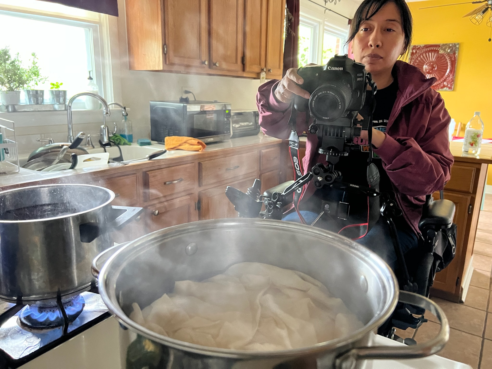 Reveca Torres is a Mexican-American female filmmaker, seen here filming in a kitchen. She is wearing a maroon sweatshirt, and operating a camera on her wheelchair. Image courtesy of Reveca Torres.