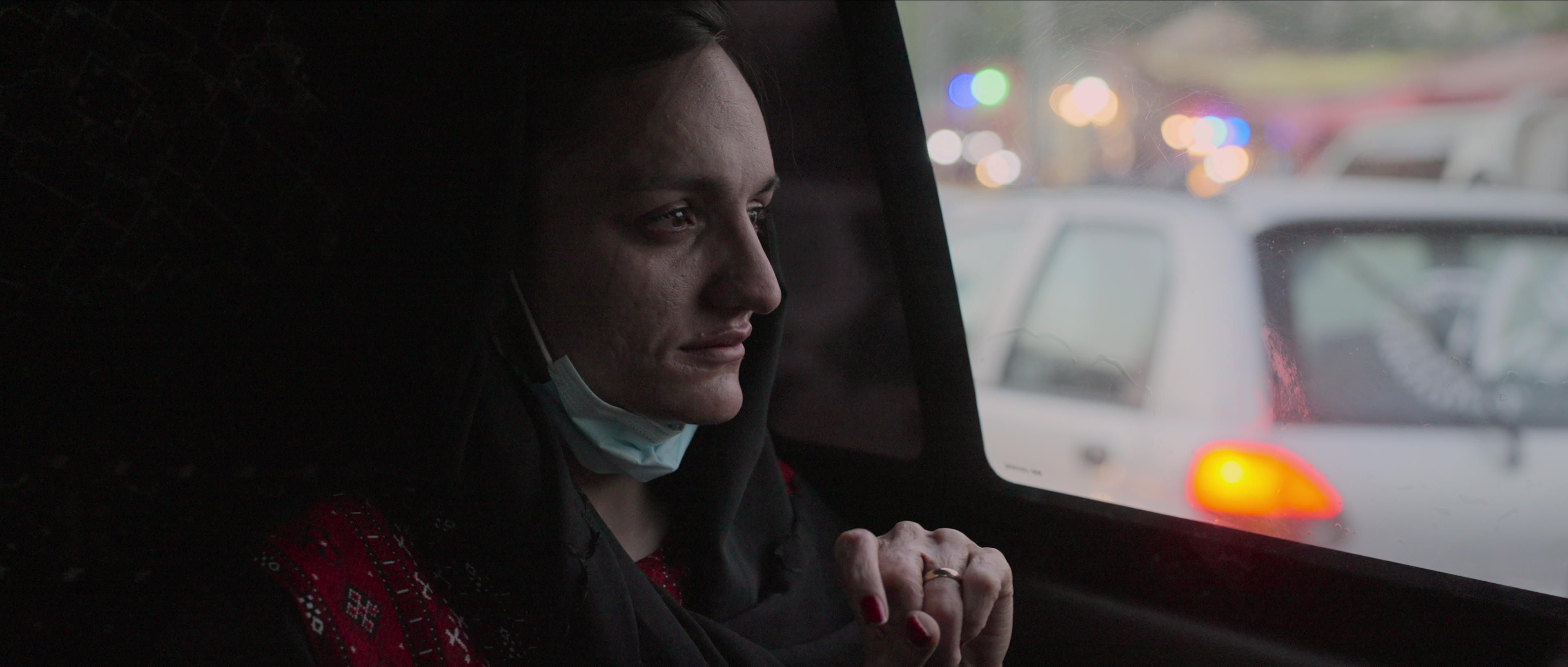 Zarifa Ghafari, an Afghan woman with black shoulder-length hair, wearing a black coat and a red patterned scarf and a surgical mask around her neck, is sitting in the back seat of a moving car, looking out the window. From Tamana Ayazi and Marcel Mettelsiefen’s In Her Hands. Courtesy of Netflix ©2022