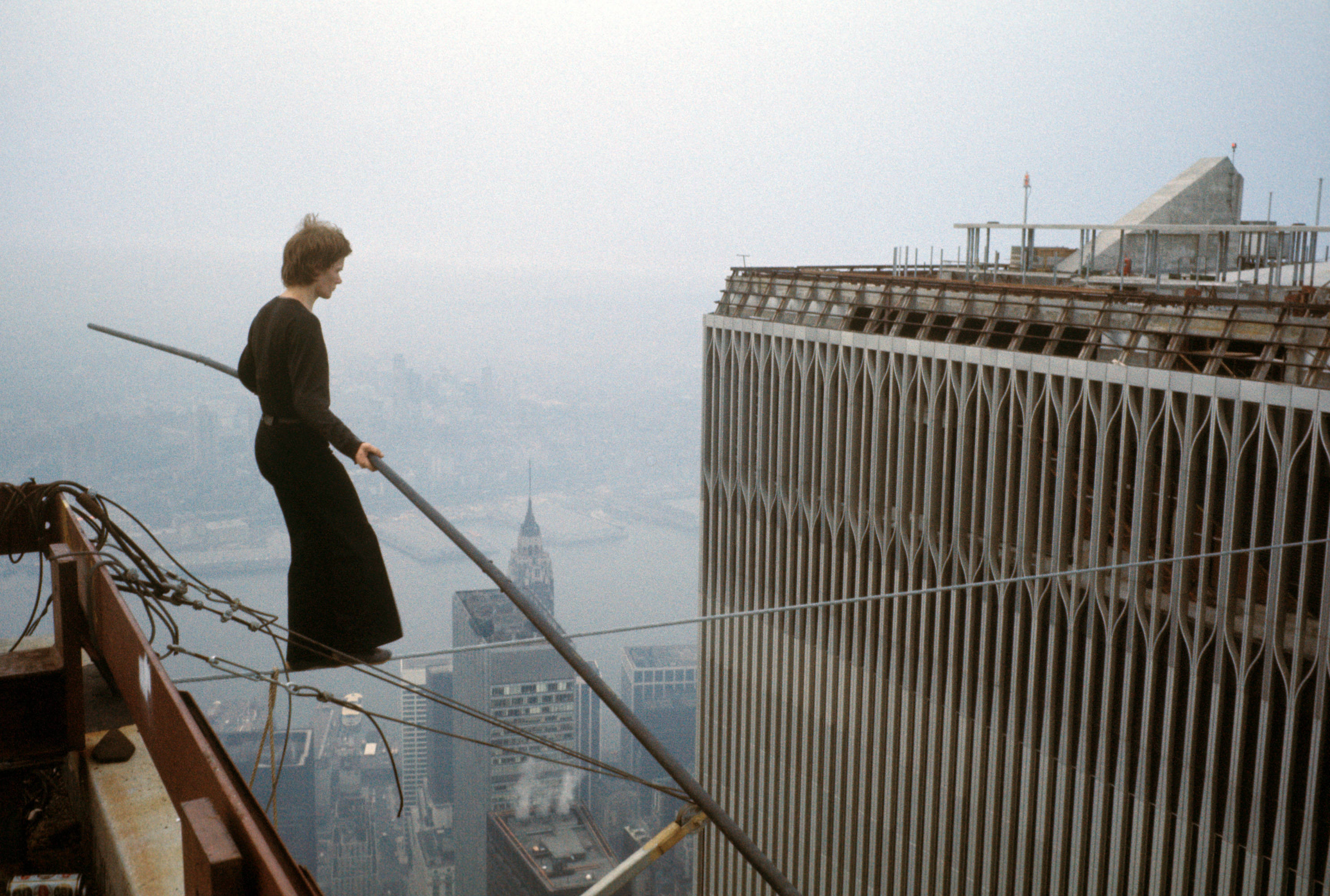 A man holding a balancing pole steps onto a high wire strung between two skyscrapers.