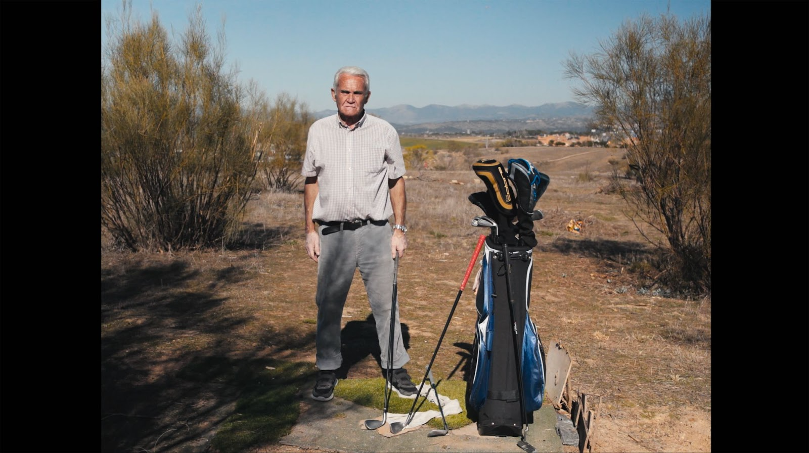 Portrait of an older man in a field, he has a golf club in his hand and next to him is the rest of the kit. He looks at the camera concentrated. The landscape is like a desert.