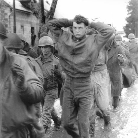 Black and white image of men in army uniforms and helmets marching, with one younger boy in plainclothes walking with his hands on his head