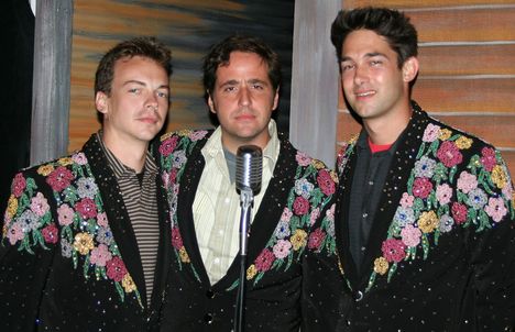 Three young men with dark hair stand around a microphone wearing black jackets with sequined flowers over their clothes