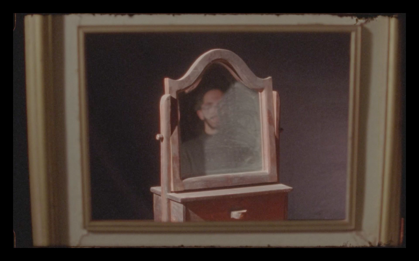 Ricardo, a pale man with glasses, dark short hair and a beard, is in the middle of the frame reflected and obscured in the mirror of a vintage vanity. The scene is framed by a window and the whole image has the soft quality of film.