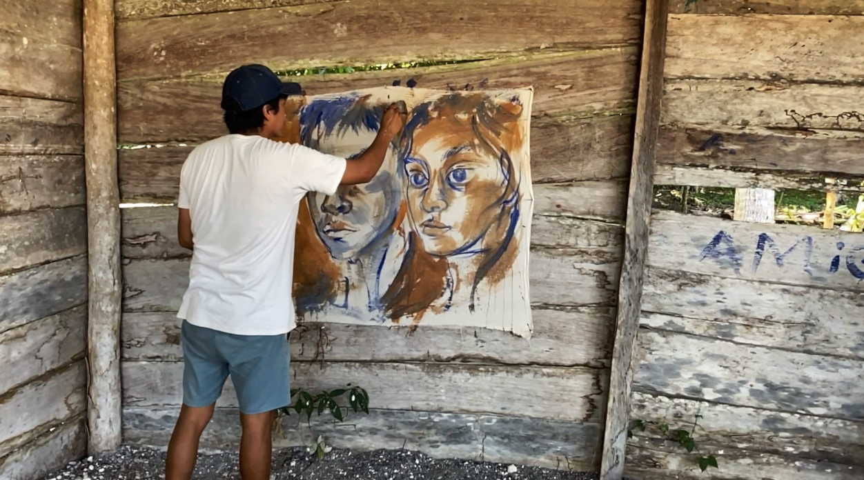 Artist Horazio Sanchez wearing a white shirt and shorts, with a blue hat on his head stands in front of a painting of two teenagers from the Calakmul Biosphere Reserve holding a paintbrush in one hand and applying it to the canvas.