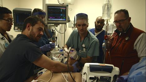 A group of man stand around a shirtless man in a hospital, all looking at monitors