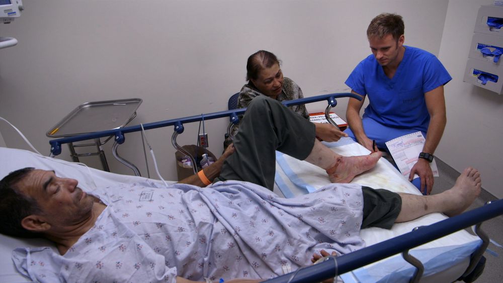 A distressed man lays on a hospital bed with a woman sitting next to him and a doctor examining his foot