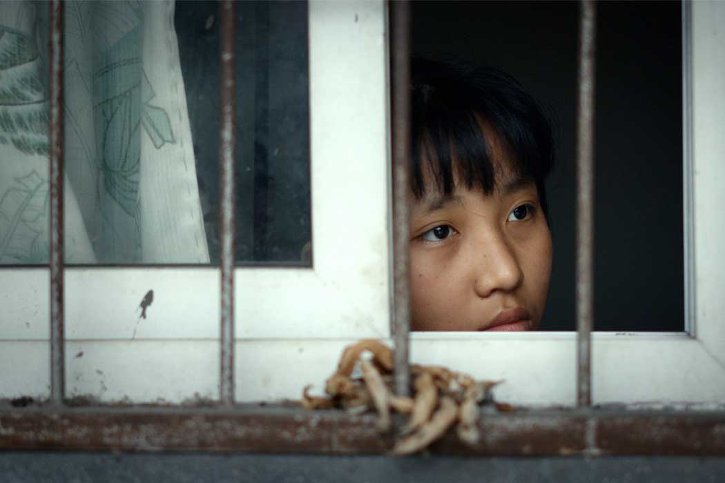 Still from 'After the Rain,' with young Chinese girl with bangs staring out of a window with a curtain and bars. Courtesy of DOC NYC.