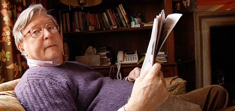 An older white man wearing glasses leans back on a couch and reads a newspaper