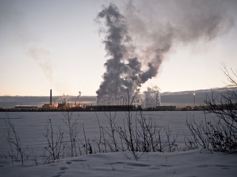 A snow covered field with a large factory in the distance with smoke billowing into the air