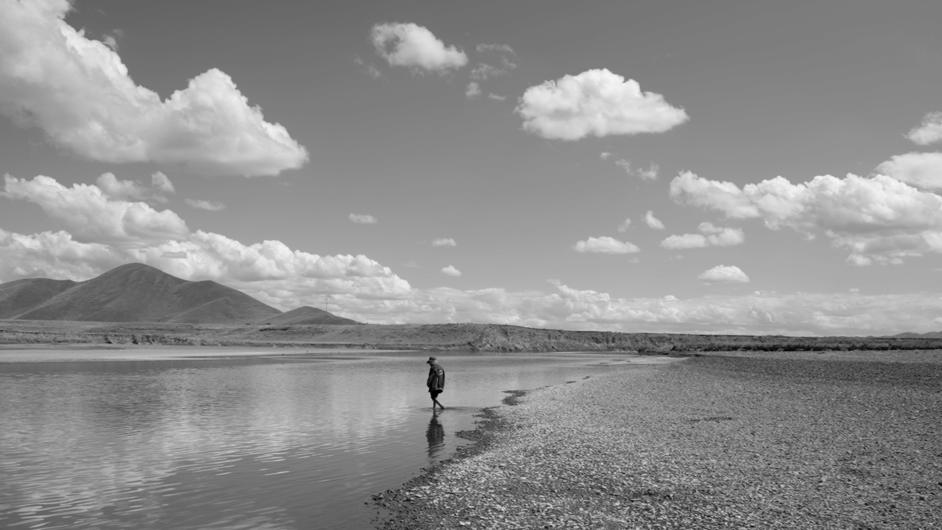 Black and white film still from 'Grietas (Cracks)' featuring a person walking on the banks of water
