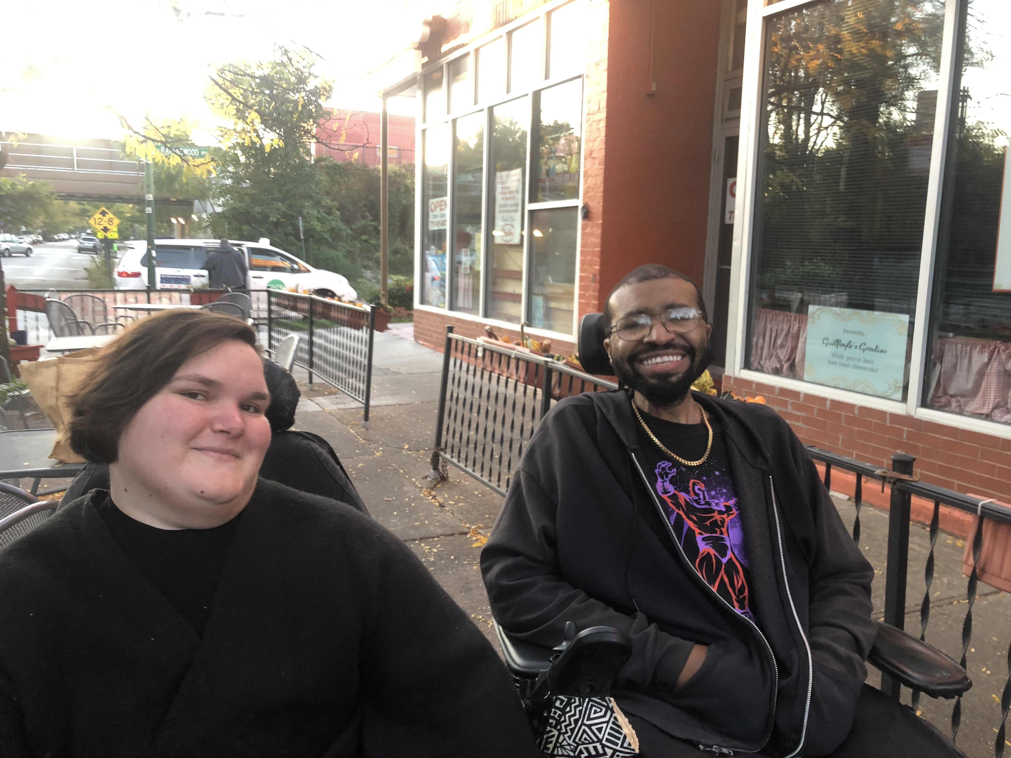 Kennedy, a white, fat, femme, and Justin, a Black man smile at the camera at an outdoor event. They both are dressed in black fall clothing and are seated in their power wheelchairs.