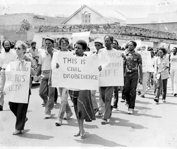 Black and white photo of protestors marching and carrying protest signs. One sign reads, "THIS IS CIVIL DISOBEDIENCE"