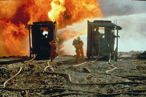 Three people in orange hazmat suits and yellow hard hats stand i a burned out field with hoses coming out of trucks, with a fire raging behind them