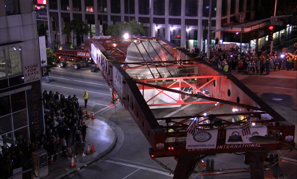 Large construction vehicles move the art installation through the street, as crowds on the sidewalk look on