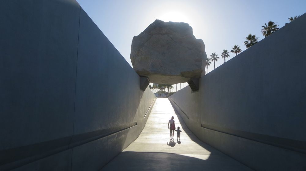 An adult holding a child's hand in the distance, walking under a large rock braced over two walls