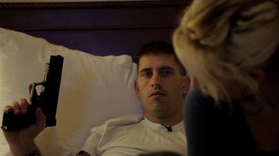 A young man lays back in a bed holding up a gun, with the profile of a blonde woman in the foreground