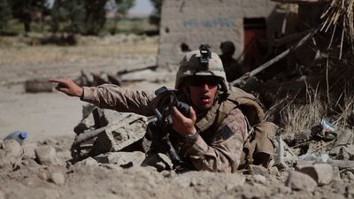 A soldier lays on the sandy ground wearing his combat helmet sunglasses, speaking into a walkie talkie and pointing to the side