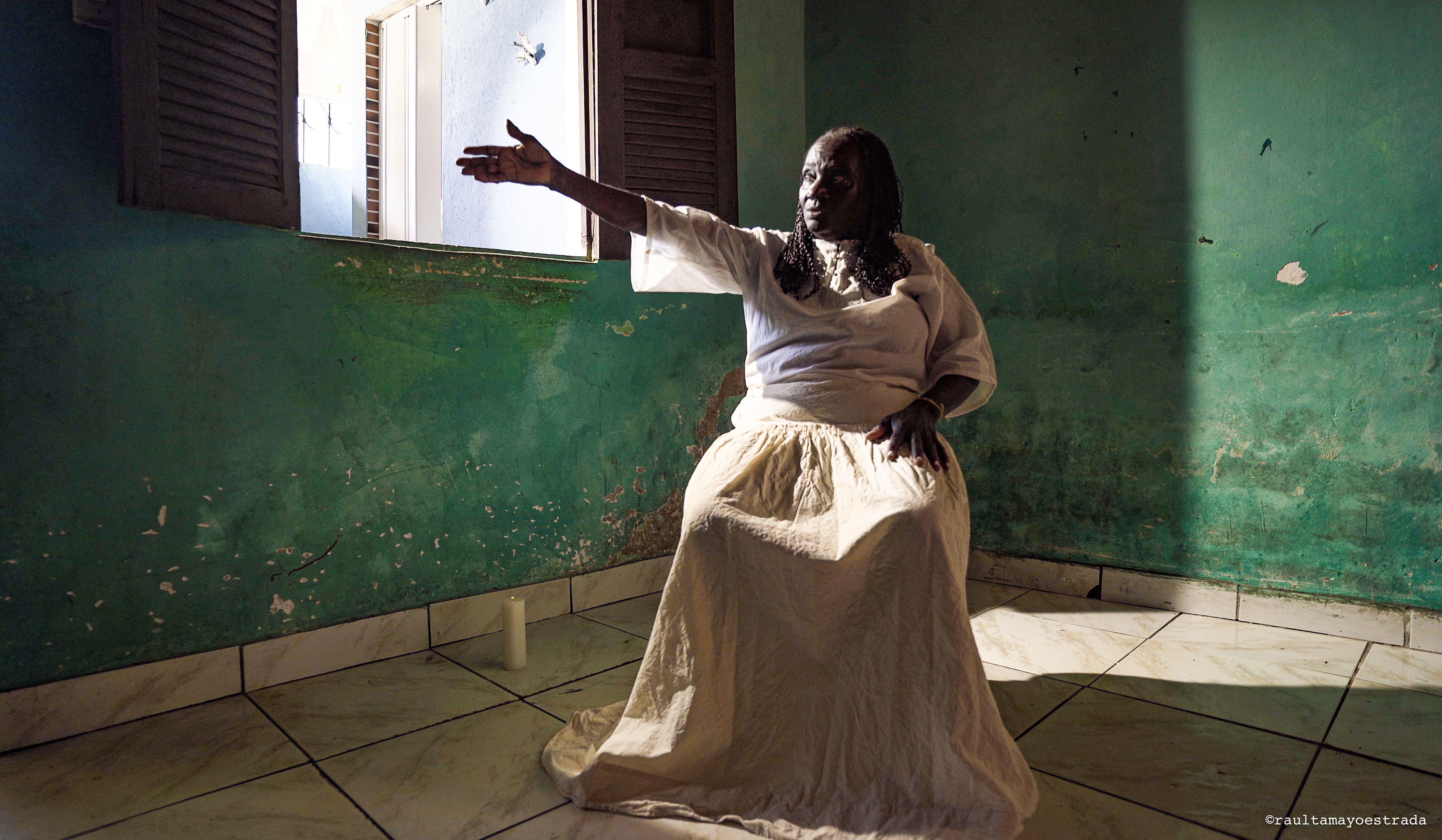 FIlm still from 'Anna Borges do Sacramento' featuring a woman with dark colored skin sitting in a chair in a room with green walls