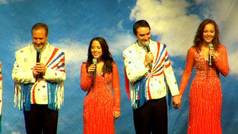 Two young women in red sparkly floor length gowns and two men in white jackets with red, white and blue sparkles and fringe stand on a stage holding microphones. One of the men and one of the women are holding hands