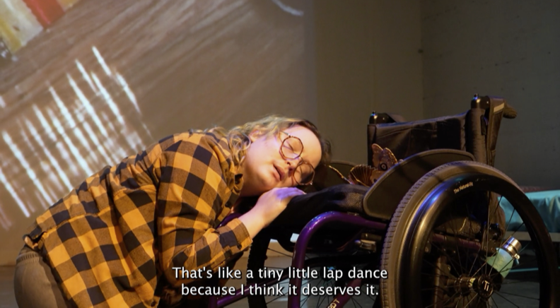 Image of a light-skinned person on a stage in front of a screen. The person is kneeling on the floor, is wearing a black and yellow checkered flannel, and glasses. They are resting their head and right hand on their black wheelchair. At the bottom of the image there is a text in white that says, “That’s like a tiny little lap dance because I think it deserves it.”