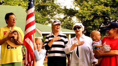 A man and two women hold their hands over their heart with their mouths open. A young girl stands between them holding a flagpole with an American flag on it. A third adult woman stands with them holding a baby