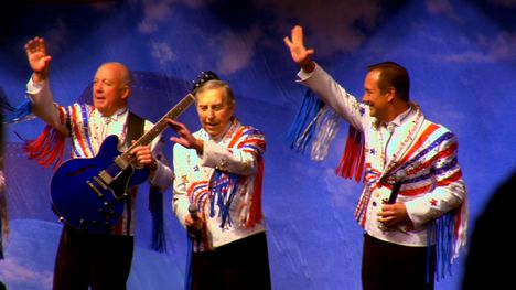 Three older men wearing white jackets with red, white and blue sparkles and fringe stand on a stage waving to a crowd. One man has a guitar and the other two are holding microphones.