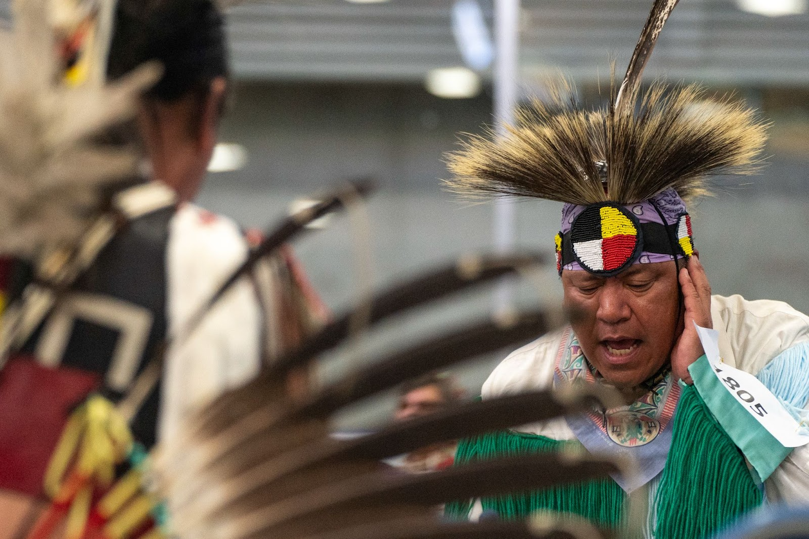 A man on the right is wearing an elaborate white, red, yellow, black, and purple beaded headdress while drumming at a ceremony at the Trout Lake Pow Wow. A blurry image of another man dressed in traditional attire with his back turned to his left.