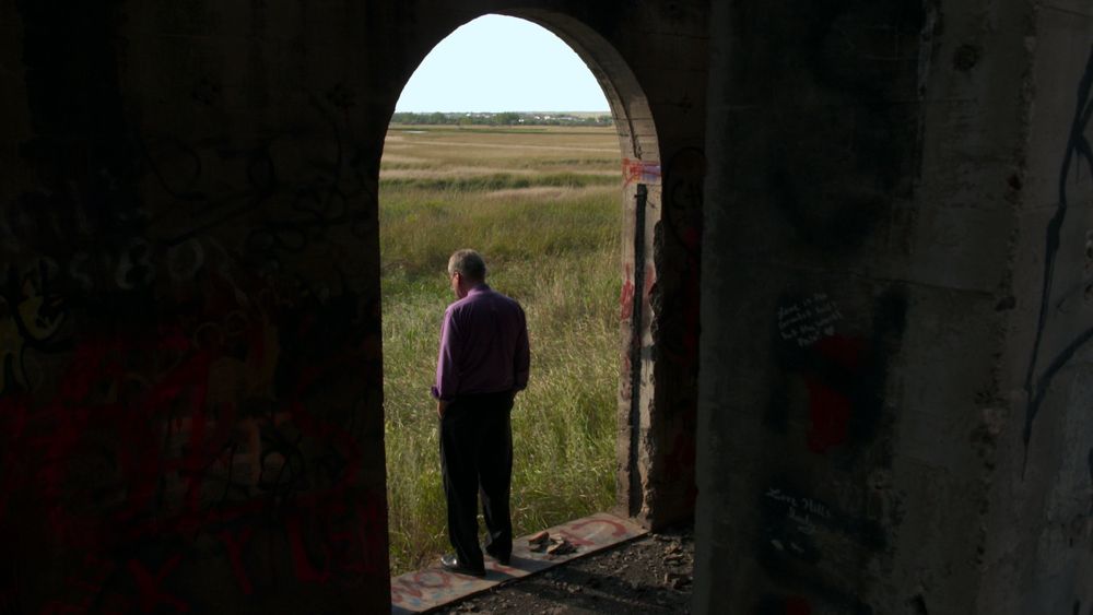 Photo of Jay Reinke from behind, standing in a large open window of an abandoned building looking out at a field of grass