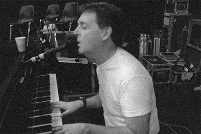 Black and white photo of Paul McCartney, a middle-aged white man, sitting at the piano singing into a microphone