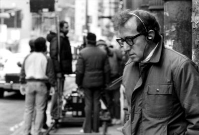 Black and white image of Woody Allen on the street, wearing headphones, glasses, and a jacket