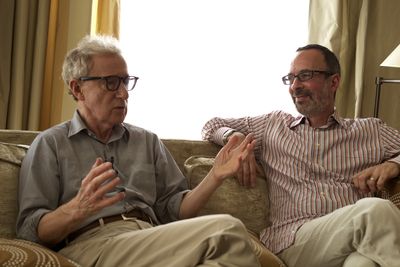 Two older white men sit on a couch in conversation. Both have glasses, button down shirts, and khakis on.