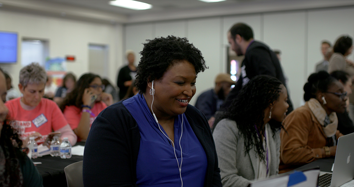 Stacy Abrams, a middle age black woman with curly hair, sits at a laptop smiling at the screen while wearing headphones