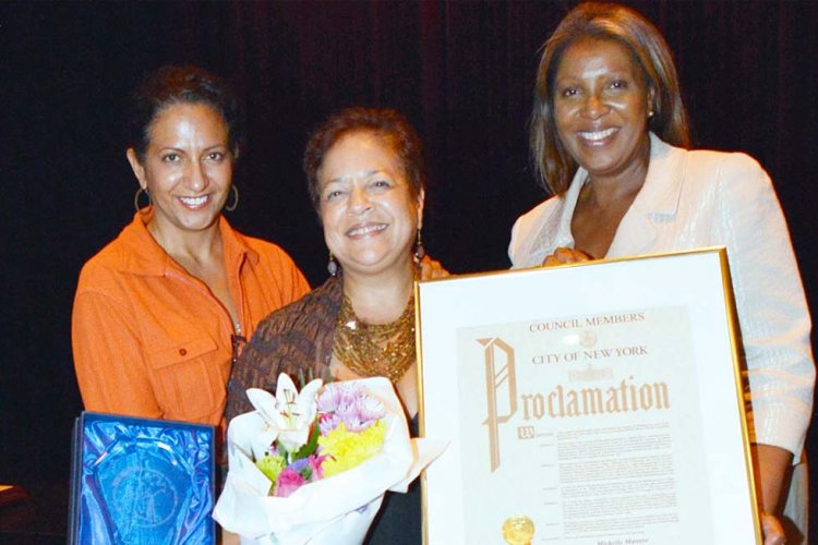 Three middle-aged women standing, one holding a bouquet of flowers and one presenting a plaque