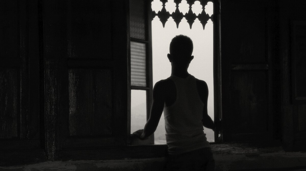 A black and white image of a silhouette of a boy sitting in a window looking out