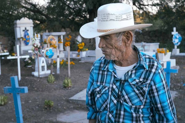 An older Mexican man wearing a blue plaid shirt and a white hat stands in a graveyard