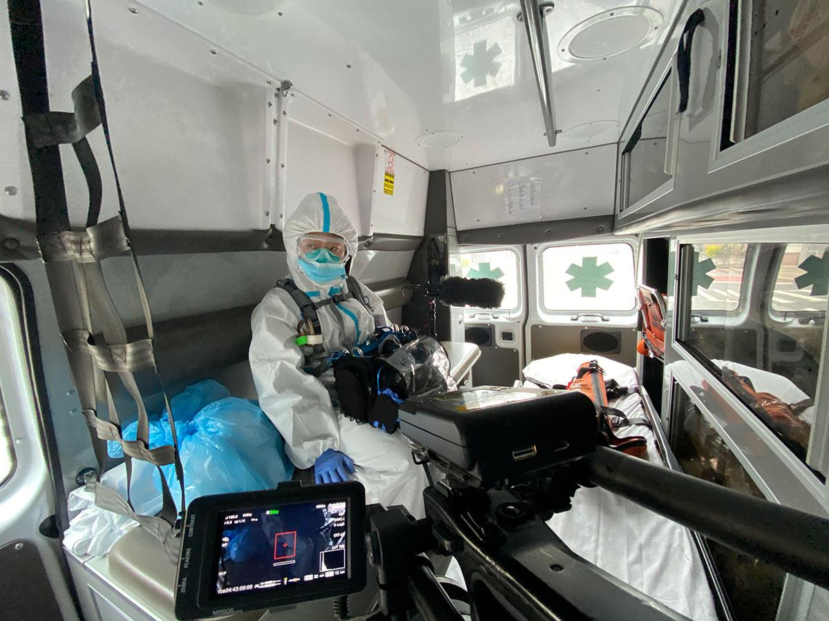 Woman in a white medical suit, mask, and goggles sits in a van and looks at a tablet