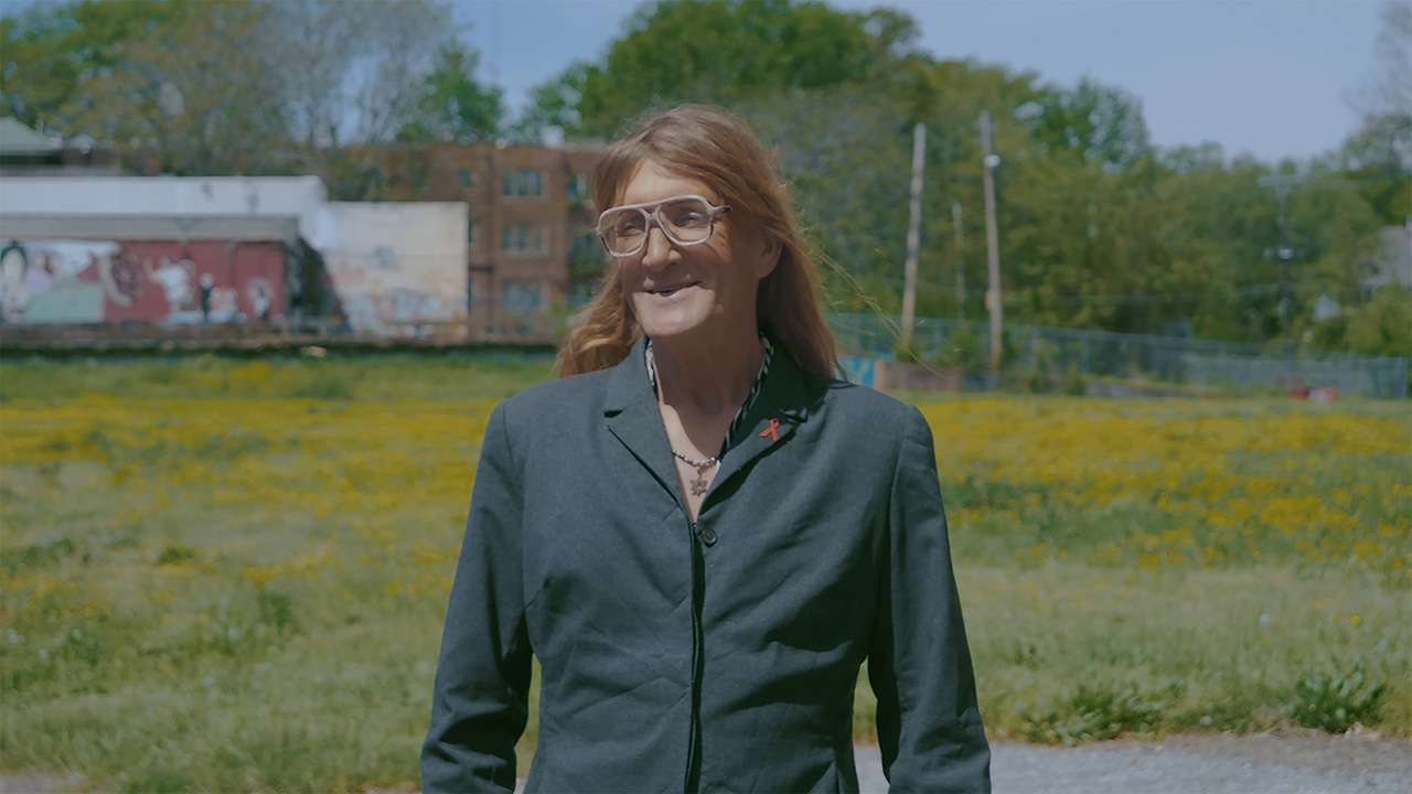 A white woman, wearing glasses and a dark green button-down shirt, with shoulder-length strawberry blond hair, stands in front of an empty lot. From Kriss Li’s ‘First Months of Freedom.’ Courtesy of DOC NYC