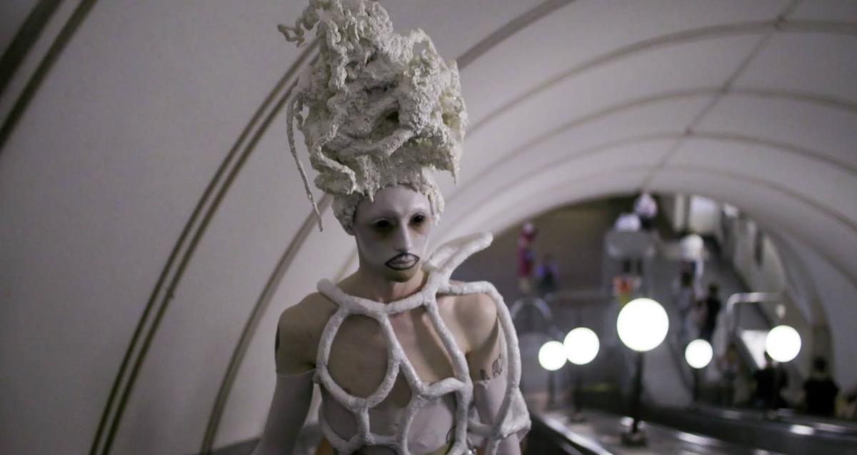 Film still from Queendom. Person riding an escalator underground wearing a large white wig, top made out of foam like material with large holes, and white, sheer gloves. The person has white and black makeup on their face. 