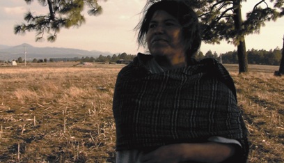 A middle aged woman stands in a field, arms crossed, looking into the distance to the side
