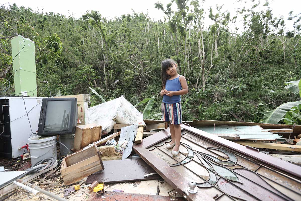 A young girl with long brown hair stands on a pile of ruins from a house. She is wearing a blue tank top and striped shorts.